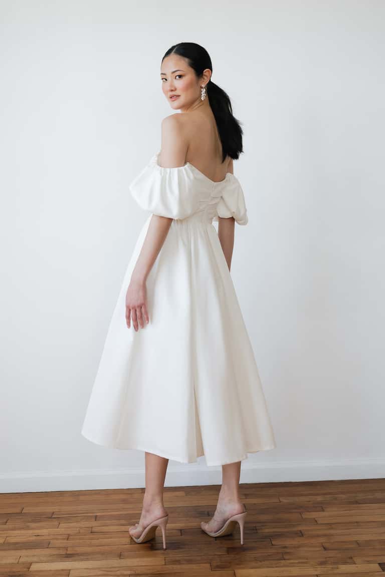Woman wearing a white off-shoulder dress against a plain white wall.