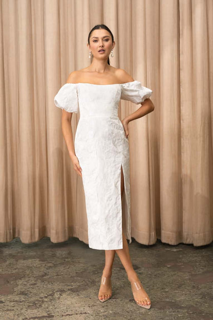 Woman wearing a white off-shoulder dress with puff sleeves against a beige curtain background