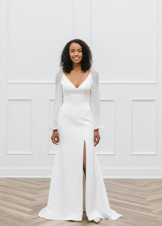 Woman wearing a white long-sleeve wedding dress with a slit on a wooden floor and white paneled wall background.