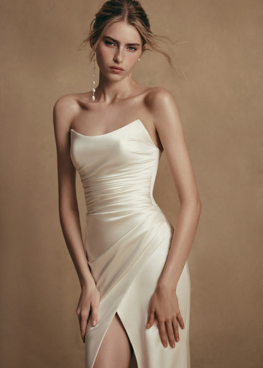 A woman modeling a strapless ivory satin gown with a ruched bodice, draped skirt, and high slit, posed against a warm beige studio backdrop.