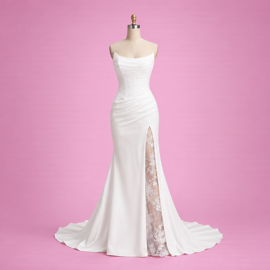 A white strapless mermaid-style evening gown displayed on a dress form against a solid pink studio background. The dress has a fitted bodice, a high slit with sheer nude lace lining, and a slight train.