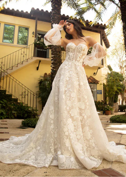 A woman wearing a voluminous ivory lace wedding gown with sheer balloon sleeves, standing outdoors in front of a yellow building with palm trees.