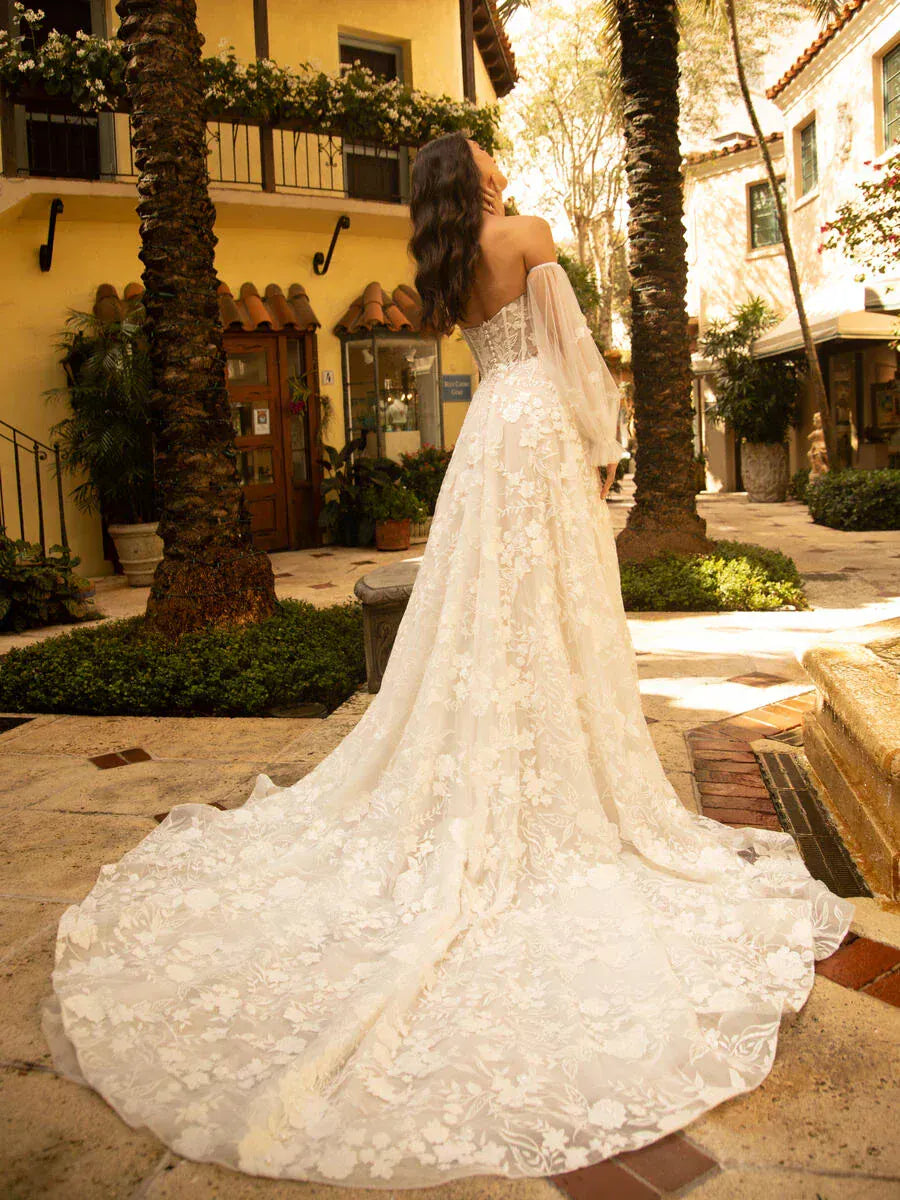 Woman in a white lace wedding dress standing in an outdoor setting with palm trees and buildings.
