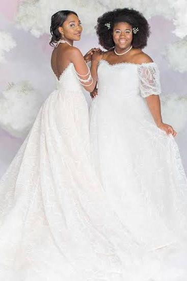 Two young women wearing white off-the-shoulder wedding dresses pose together against a pastel pink and white cloud-patterned backdrop.
