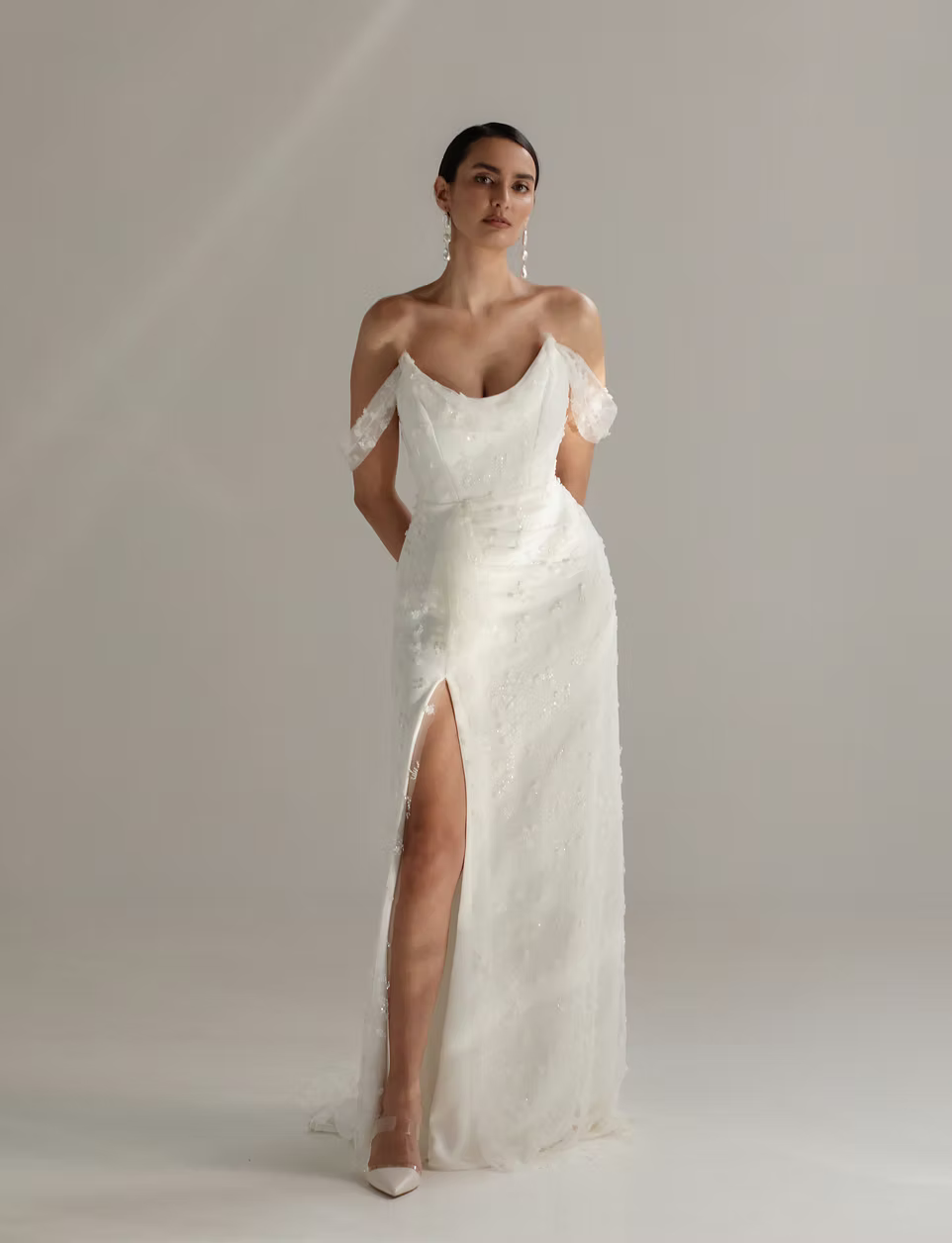 A woman modeling a floor-length off-the-shoulder white gown with a deep V neckline, sheer lace flutter sleeves, a fitted waist, and a high front slit, photographed against a soft light gray studio background.