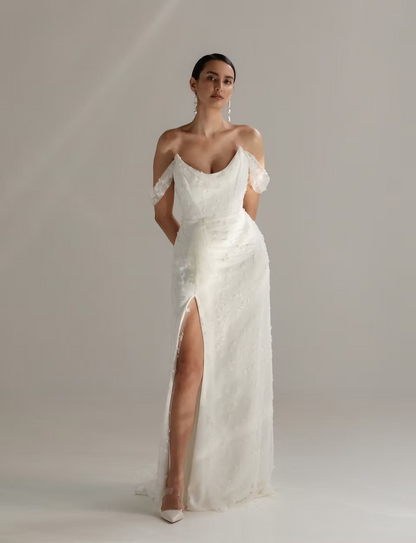 A woman modeling a floor-length off-the-shoulder white gown with a deep V neckline, sheer lace flutter sleeves, a fitted waist, and a high front slit, photographed against a soft light gray studio background.