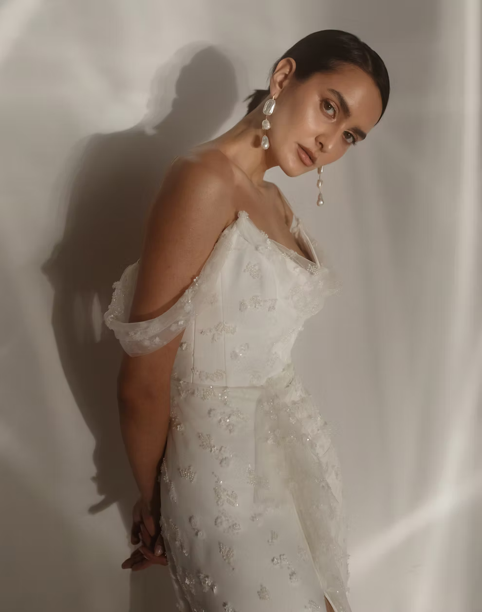A woman wearing a white off-the-shoulder embellished dress, posing against a draped white fabric backdrop with dramatic shadows.