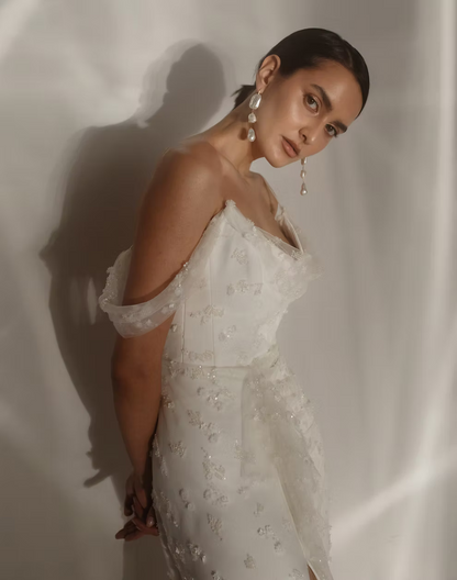 A woman wearing a white off-the-shoulder embellished dress, posing against a draped white fabric backdrop with dramatic shadows.