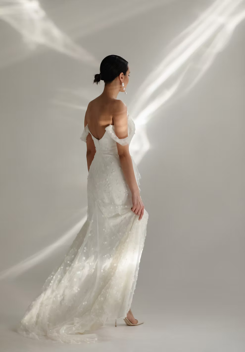 A woman wearing a white lace wedding gown stands with her back to the camera in a studio with dramatic light beams on a light gray background.