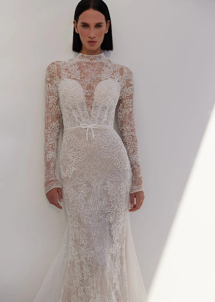 An intriguing and seductive, stylish and alluring mermaid silhouette dress in lightskin color, made of milky beaded lace with a Chantilly effect, will suit a confident bride. The corset has a deep heart-shaped neckline. An embroidered lace tank top with a stand-up collar and long sleeves is sewn on top of the corset. The back of the dress is made of skillfully embroidered lace. The back is fastened with loops and buttons. The skirt is translucent, close-fitting in the hip area and has an elegant train. The 