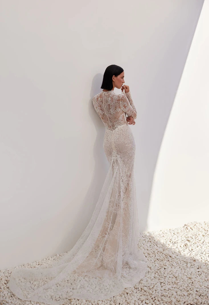 An intriguing and seductive, stylish and alluring mermaid silhouette dress in lightskin color, made of milky beaded lace with a Chantilly effect, will suit a confident bride. The corset has a deep heart-shaped neckline. An embroidered lace tank top with a stand-up collar and long sleeves is sewn on top of the corset. The back of the dress is made of skillfully embroidered lace. The back is fastened with loops and buttons. The skirt is translucent, close-fitting in the hip area and has an elegant train. The 