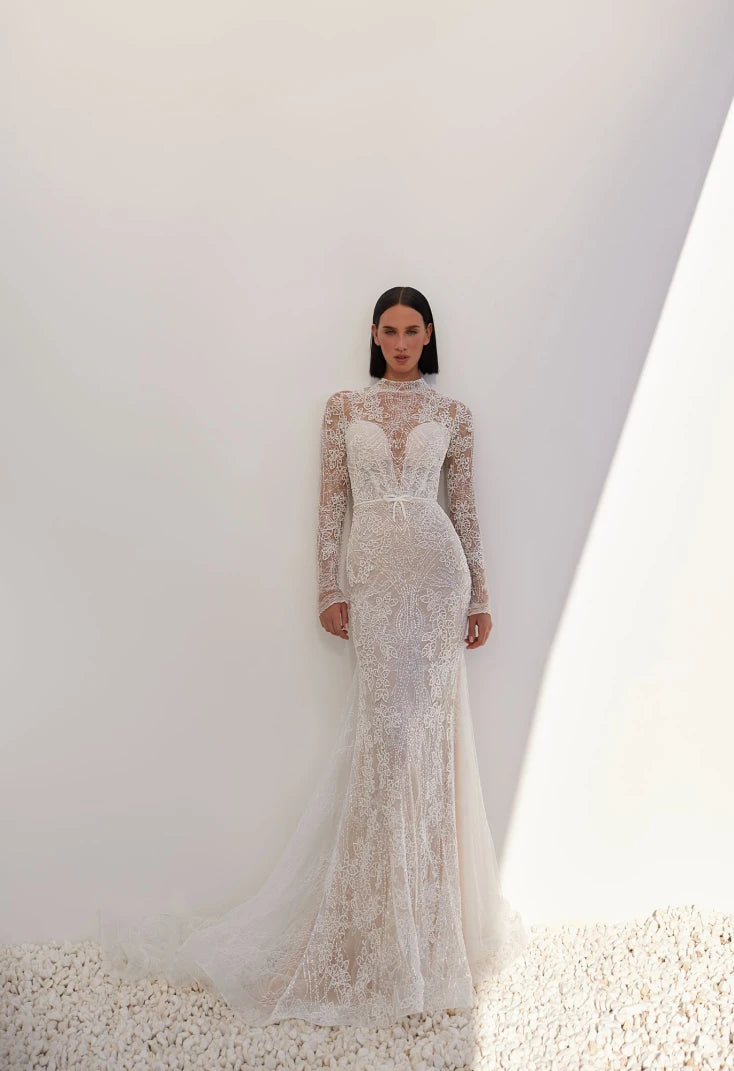 An intriguing and seductive, stylish and alluring mermaid silhouette dress in lightskin color, made of milky beaded lace with a Chantilly effect, will suit a confident bride. The corset has a deep heart-shaped neckline. An embroidered lace tank top with a stand-up collar and long sleeves is sewn on top of the corset. The back of the dress is made of skillfully embroidered lace. The back is fastened with loops and buttons. The skirt is translucent, close-fitting in the hip area and has an elegant train. The 