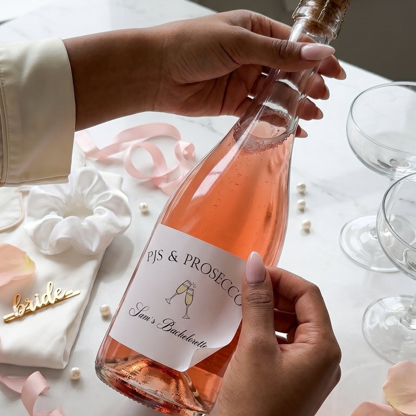 Bride holding a bottle of Prosecco with a customized bachelorette label. She is adhering the stick label that reads, "pjs & processco, sam's bachelorette" the background is a light pink and white decor is bride themed elements.