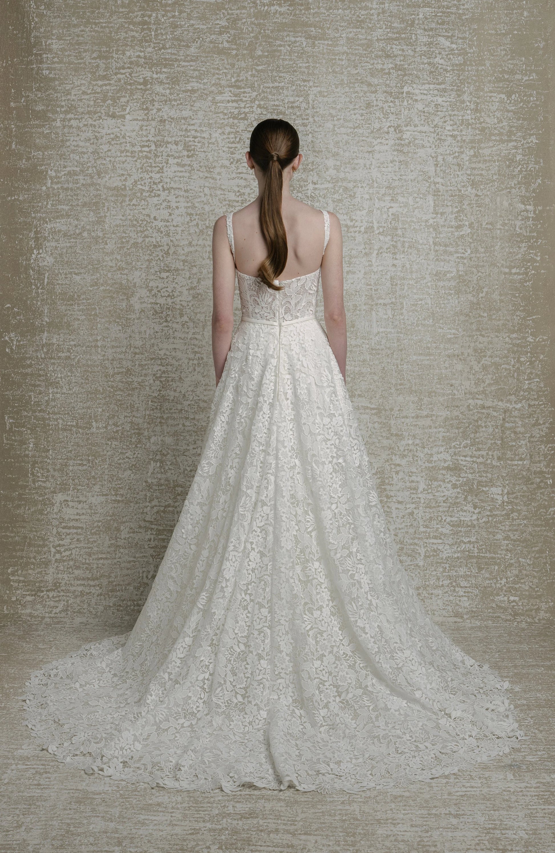 Woman wearing a white lace wedding dress against a textured beige wall.