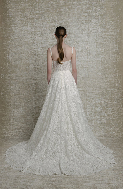 Woman wearing a white lace wedding dress against a textured beige wall.