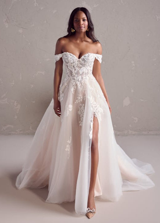 Woman wearing a white off-shoulder lace wedding dress with a high slit on a plain background
