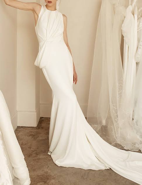 A woman models a sleeveless white wedding gown with a high neckline, gathered waist, and a long flowing train in an indoor bridal shop setting with other gowns hanging in the background.