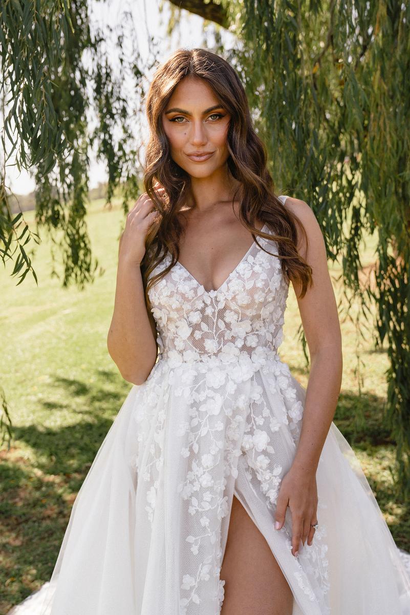 Woman in a white floral wedding dress standing outdoors with greenery.