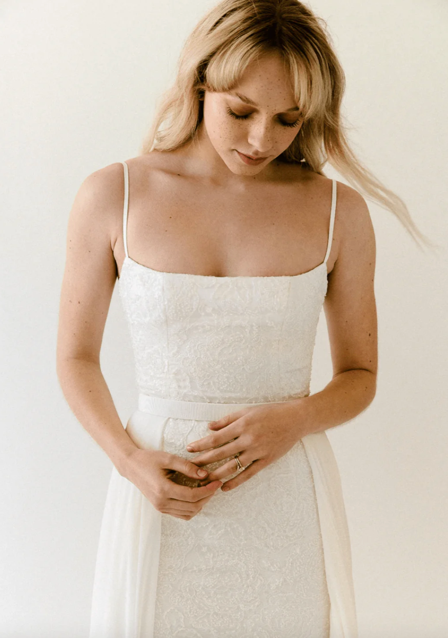 Truvelle Celine is a captivating lace slip gown with a detachable chiffon overskirt! Thin straps highlight the square neckline, while a soft train accents the column skirt. Peony-embroidered beaded lace with dazzling sequins crafts the slim silhouette of the gown.