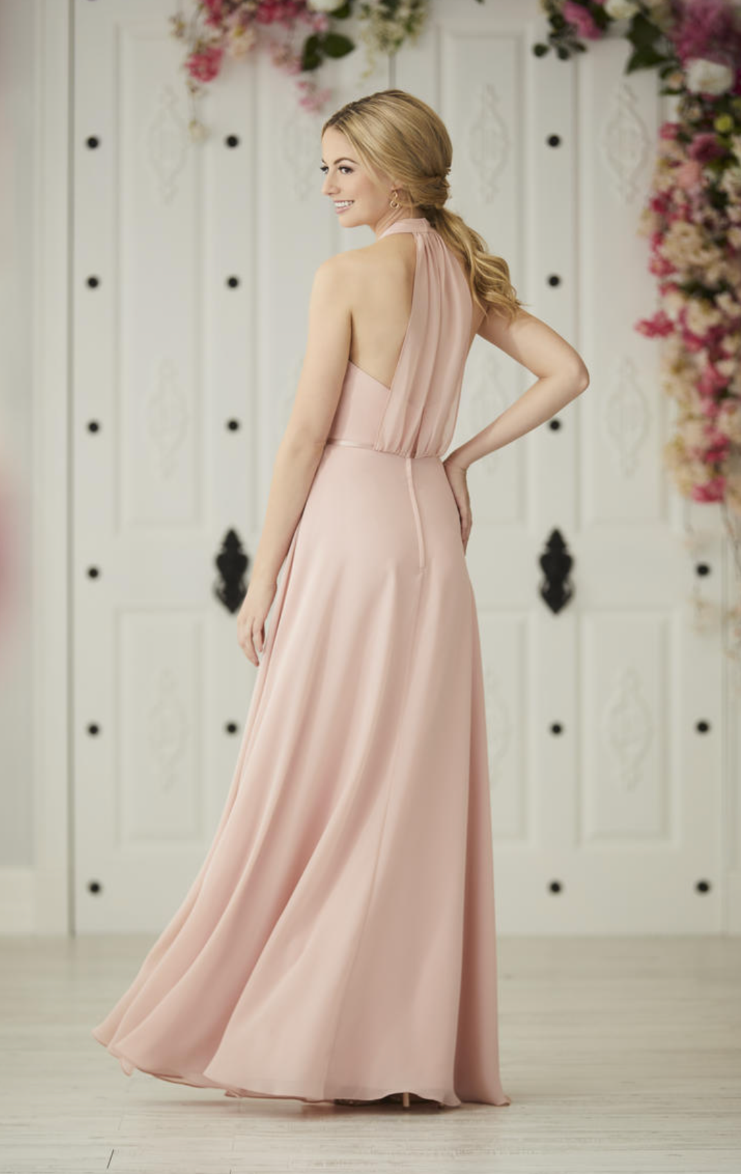 A woman modeling a floor-length blush pink gown from the back, standing in front of a white decorative door with floral arrangements on either side.