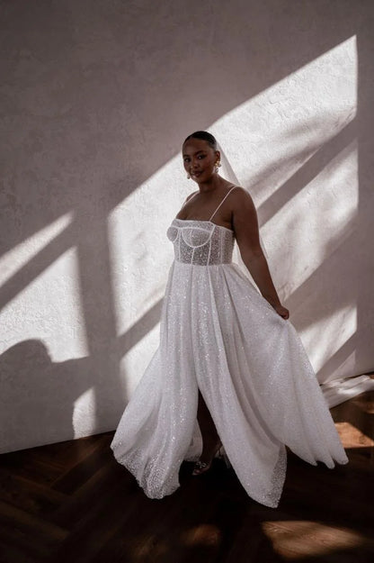 A woman wearing a white sleeveless wedding gown with a sheer corset-style bodice and a flowing textured skirt, standing in a room with dramatic diagonal sunlight and shadows on a textured wall.