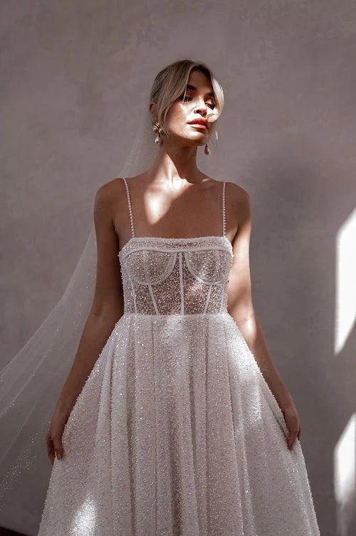 A woman wearing a sleeveless white wedding gown with a sheer lace corset bodice and a full textured tulle skirt, standing against a warm neutral wall with dramatic sunlight and shadows.