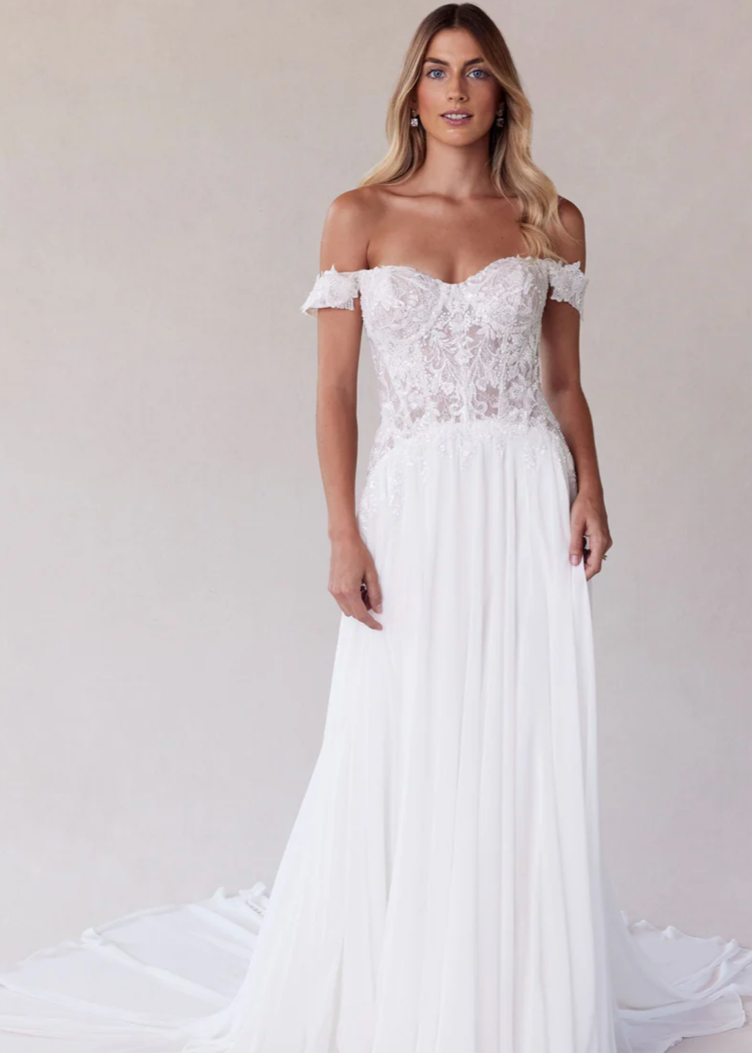 Woman wearing a white off-shoulder wedding dress with lace details on a plain background