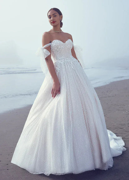 A woman wearing a voluminous off-the-shoulder white wedding gown stands on a beach with the ocean and hazy sky in the background.