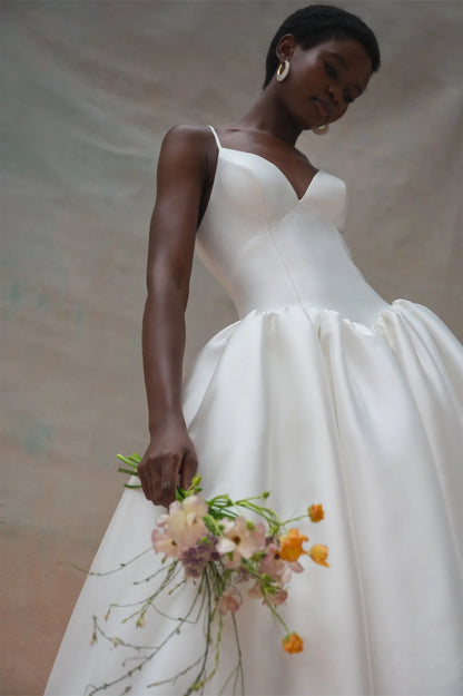 A woman wearing a white wedding dress with thin straps and a voluminous skirt, holding a small bouquet of pink, peach, and orange flowers against a soft beige studio backdrop.
