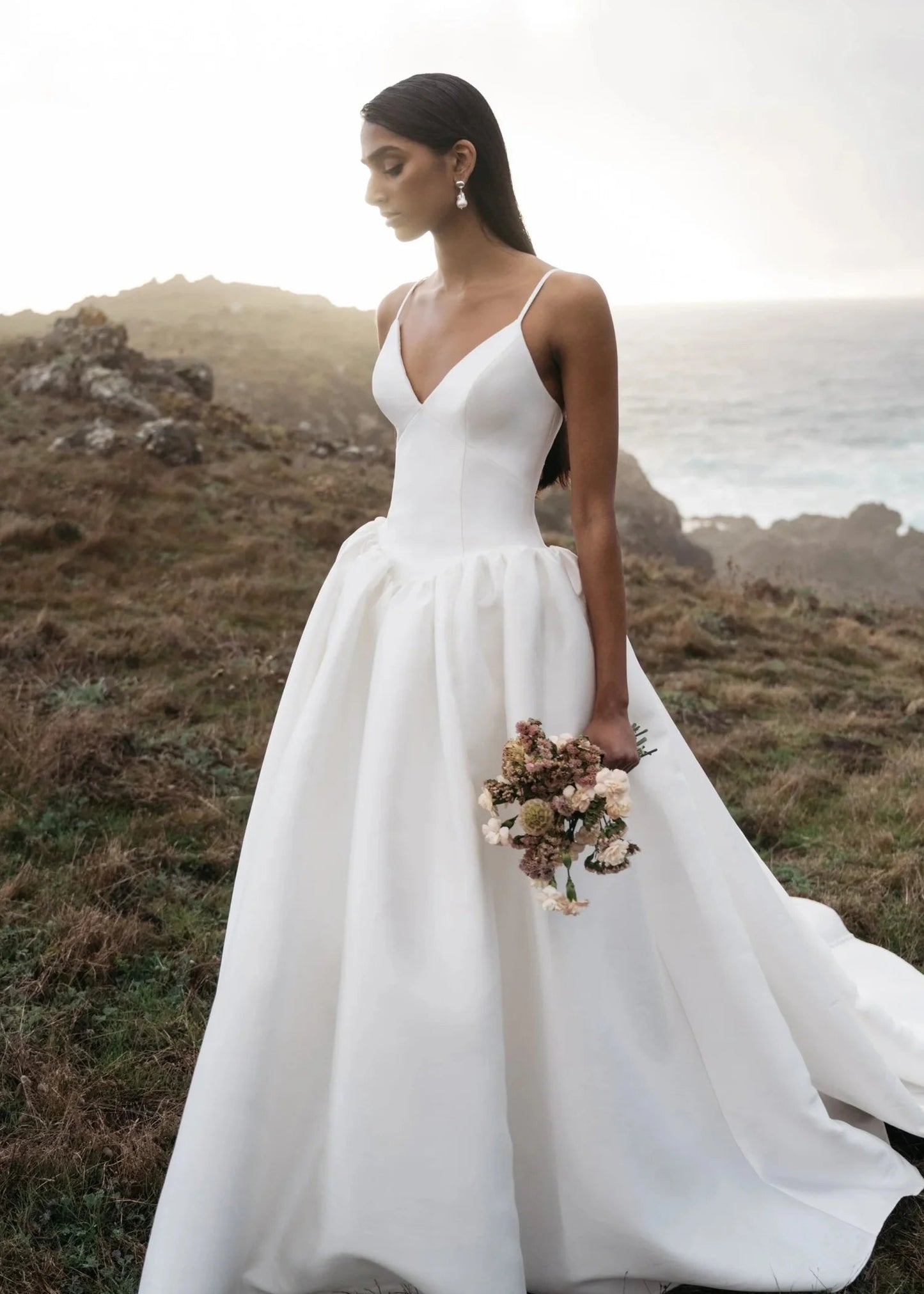 A woman wearing a white sleeveless wedding gown with a deep V-neckline and a full skirt, standing outdoors on a grassy coastal hillside and holding a bouquet of flowers.