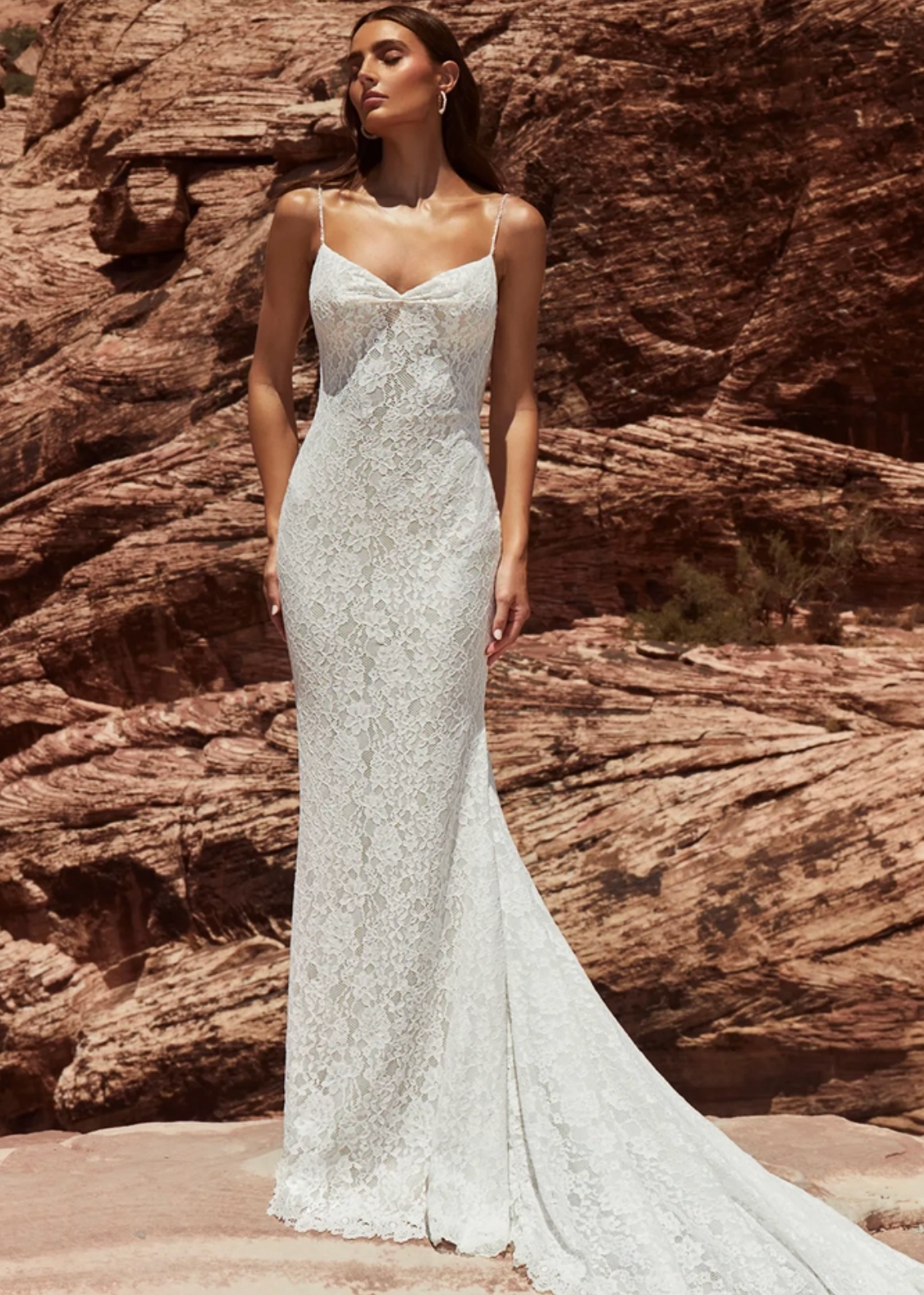 A woman wearing a fitted white lace wedding gown with thin straps and a long train, standing outdoors in front of red rock desert formations.