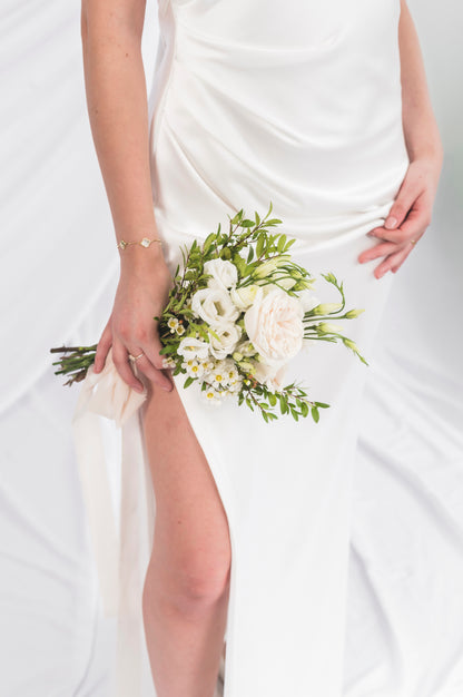A close-up of a bride wearing a white satin wedding dress with a thigh-high slit, holding a bouquet of white flowers and green leaves. She is wearing a delicate gold bracelet.