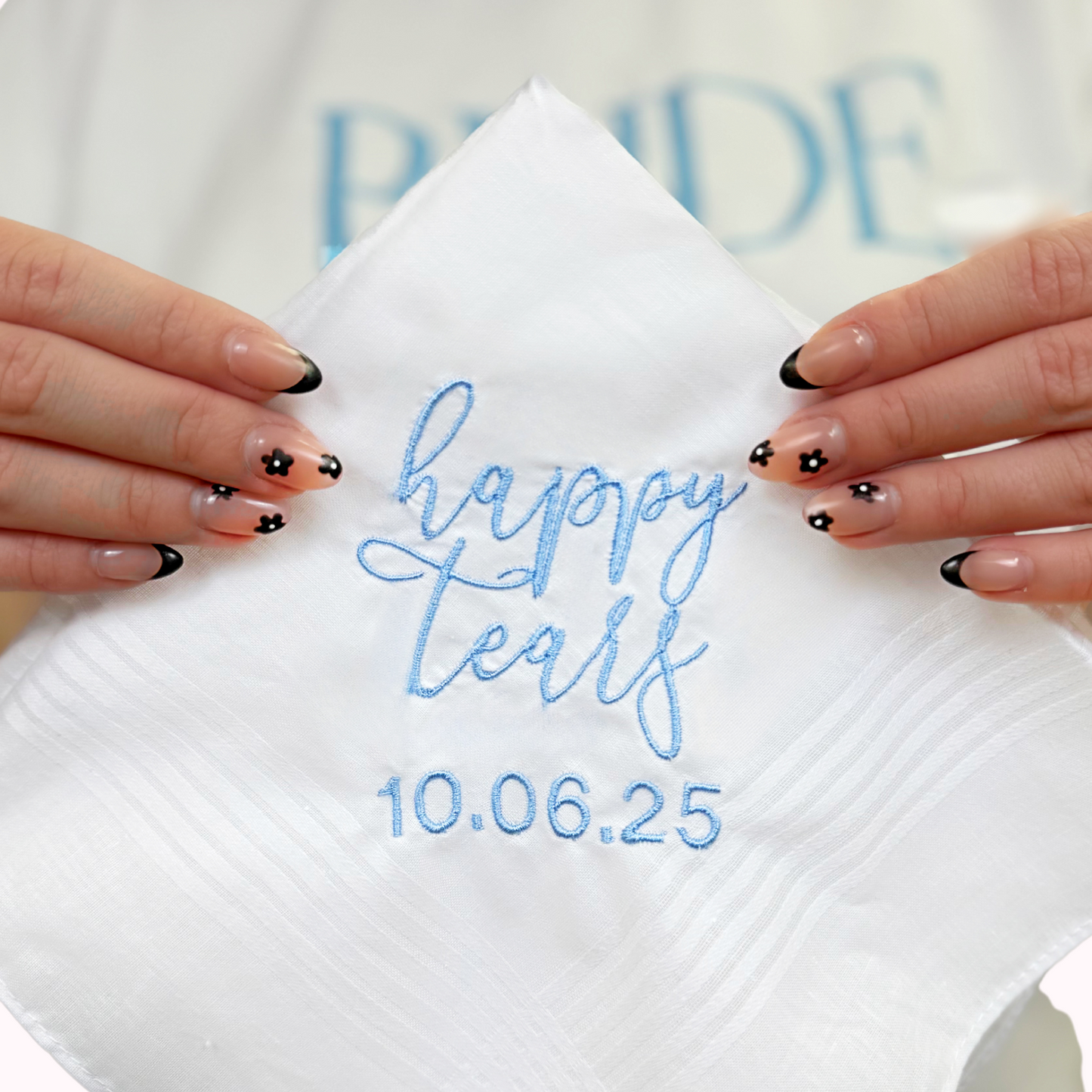 A close up of a white handkerchief with blue embroidered writing that says "happy tears 10.06.2025"