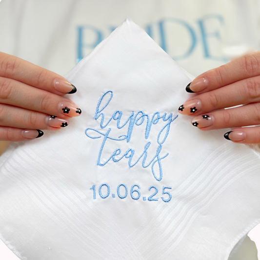 A close up of a white handkerchief with blue embroidered writing that says "happy tears 10.06.2025"