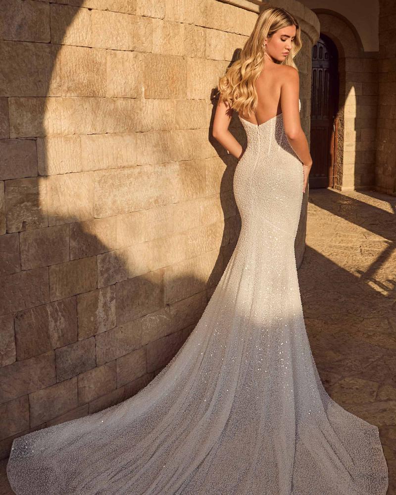 A woman wearing a sparkling silver sheath wedding gown with a backless open back and long train, posed outdoors against a sunlit stone wall. The dress is Ziva by Calla Blanch, available at NKIN