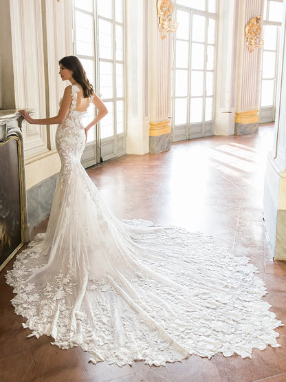 A woman wearing a fitted white lace mermaid-style wedding gown with a long dramatic train stands in a bright classical interior with tall windows and polished wood floors.Tahlia by Enzoani available at NKIN in Ottawa