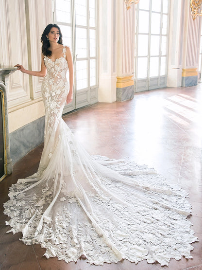 A woman models a strapless ivory floral lace mermaid wedding gown with a long sheer overskirt and dramatic train, posed in a grand sunlit palace interior with tall windows and columns. Tahlia by Enzoani available at NKIN in Ottawa