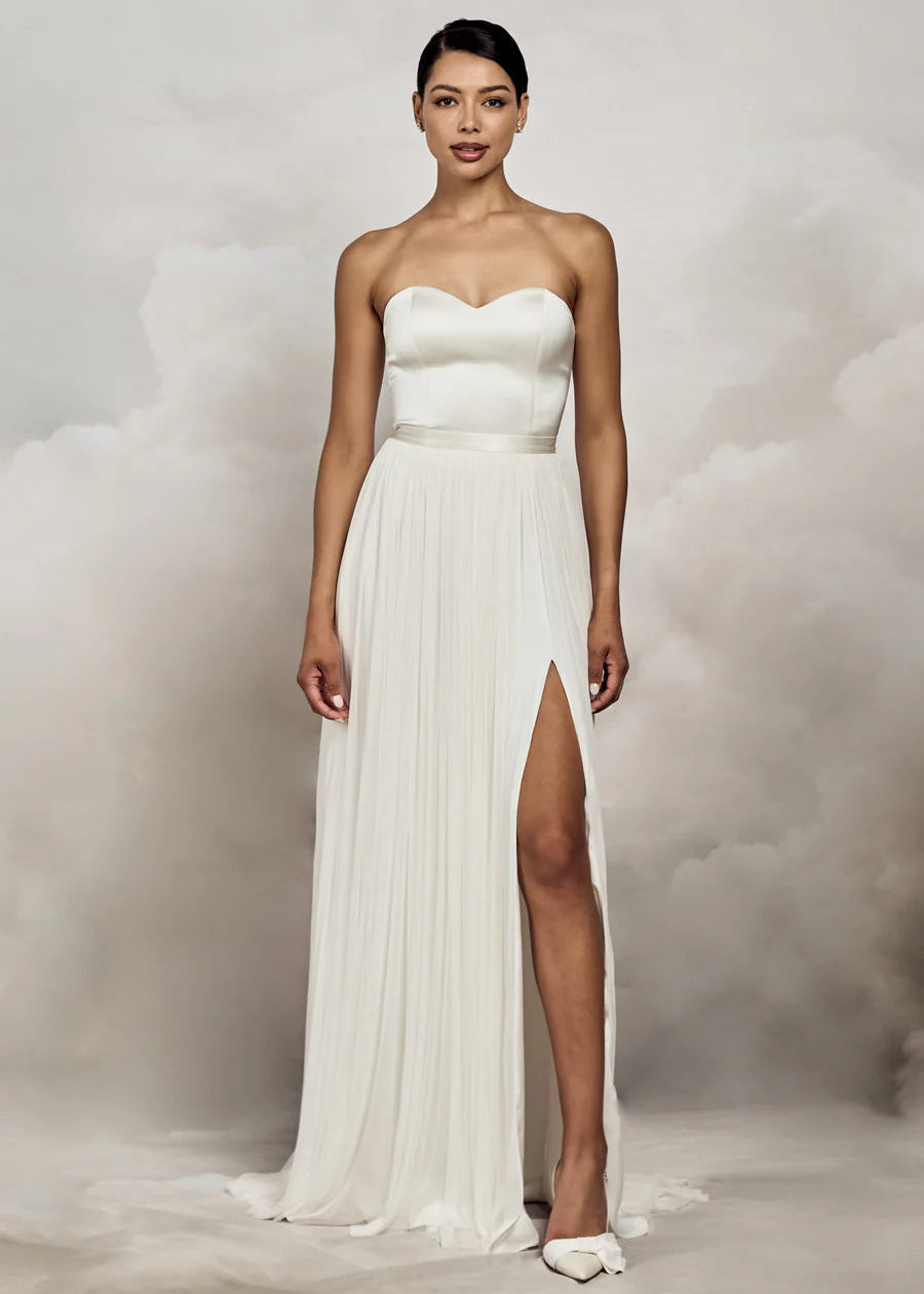 A woman modeling a strapless white wedding gown with a sweetheart neckline, fitted bodice, and a high thigh slit, photographed against a soft gray cloud-like studio backdrop.