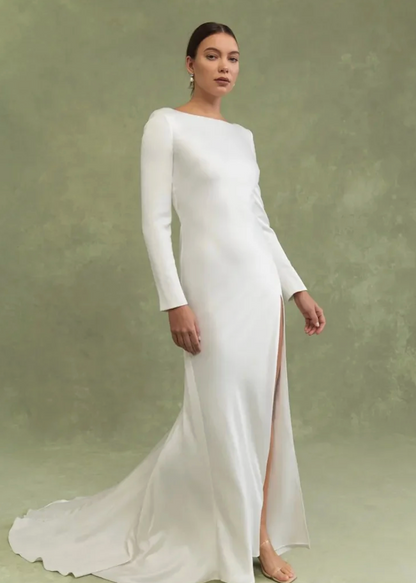 A woman models a floor-length white gown with long sleeves, a boat neckline, and a high front slit against a textured sage green studio backdrop.