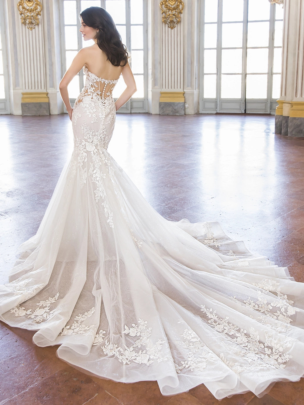 A woman wearing a strapless mermaid-style wedding gown with floral lace applique and a long sheer train, photographed in a grand interior hall with tall windows and polished wood floors.