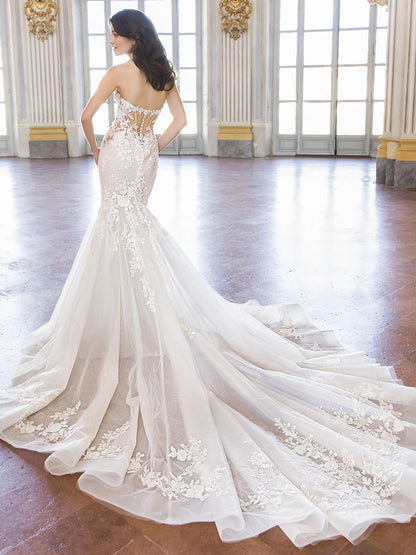 A woman wearing a strapless mermaid-style wedding gown with floral lace applique and a long sheer train, photographed in a grand interior hall with tall windows and polished wood floors.