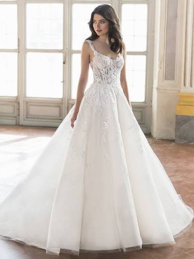 A woman wearing a white ball gown wedding dress with lace and tulle details, standing in a bright room with large windows.