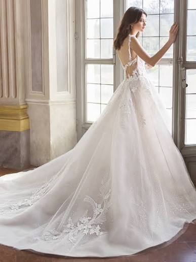 A woman wearing a white wedding gown with a long train, standing in a bright room with large windows. The dress has lace detailing and a sheer overlay.