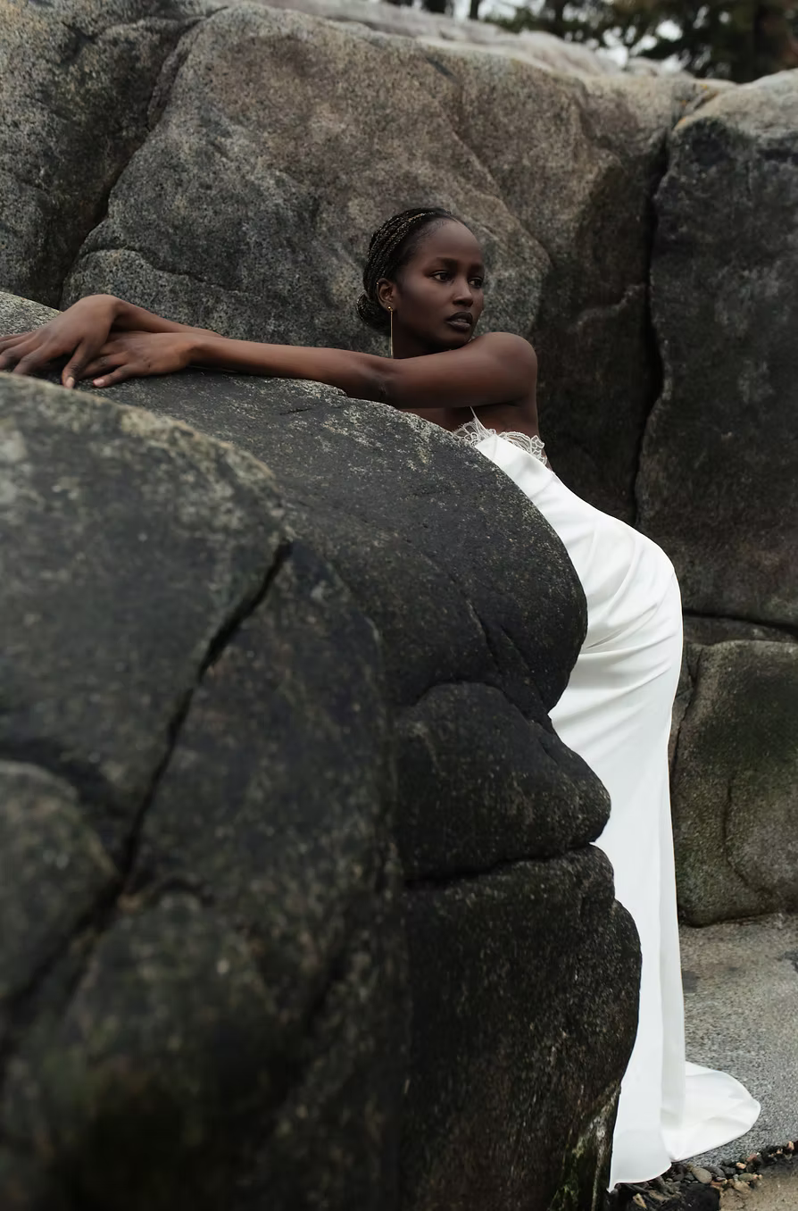 A woman in a white sleeveless gown leans against large dark rocks outdoors, with the dress pooling on the ground beside her.