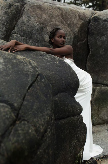 A woman in a white sleeveless gown leans against large dark rocks outdoors, with the dress pooling on the ground beside her.