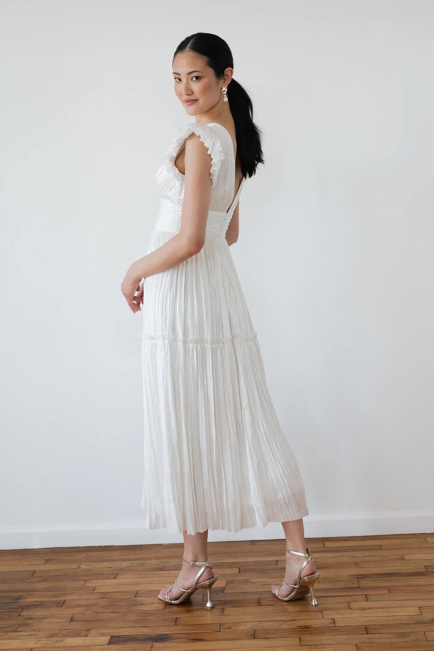 Woman wearing a white dress with cut-out shoulders and pleated skirt on a plain background