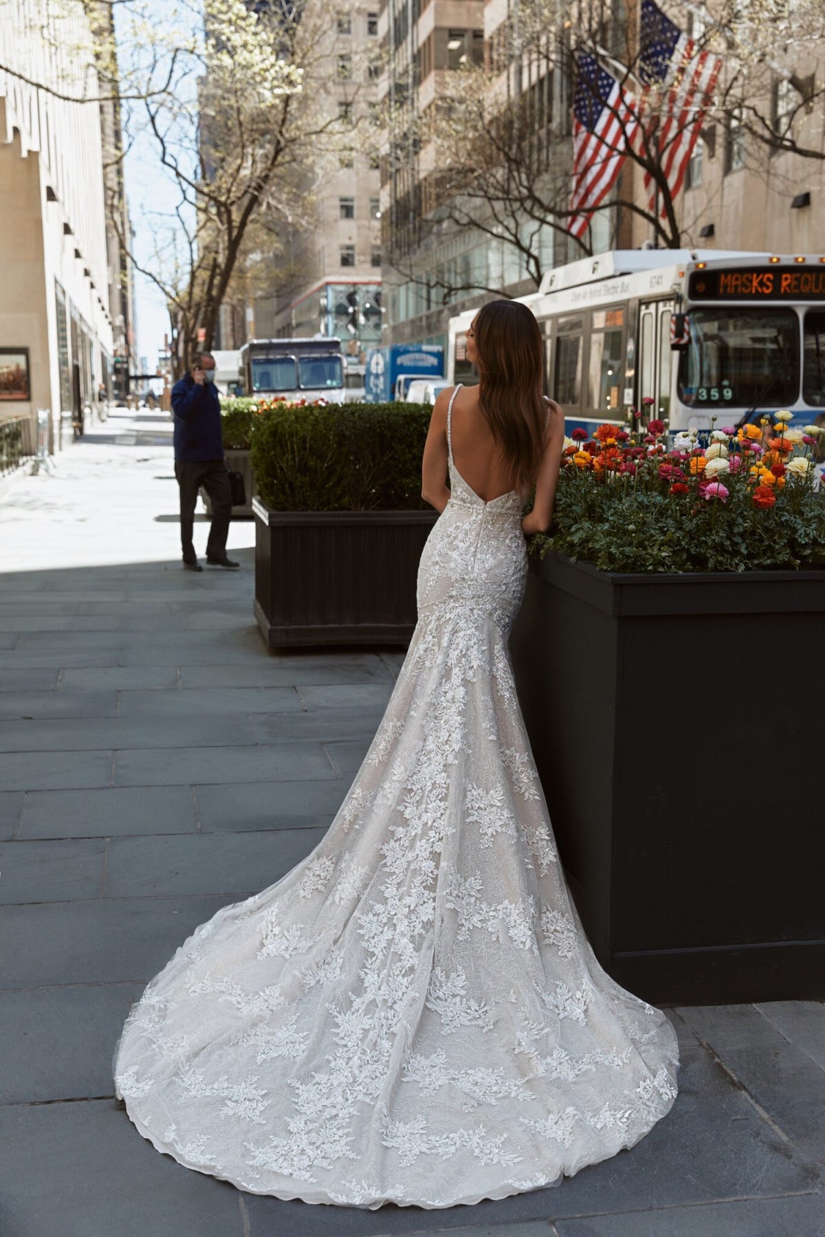 A woman wearing a white lace mermaid-style wedding gown with a long train stands on a city sidewalk, photographed from behind. The street scene includes buildings, trees, flowers, a bus, and an American flag in the background.