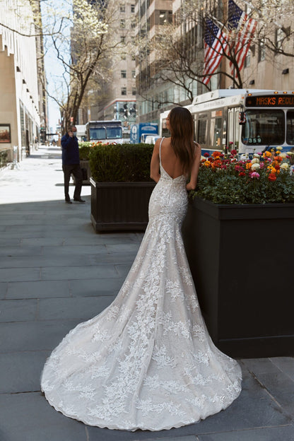 A woman wearing a white lace mermaid-style wedding gown with a long train stands on a city sidewalk, photographed from behind. The street scene includes buildings, trees, flowers, a bus, and an American flag in the background.