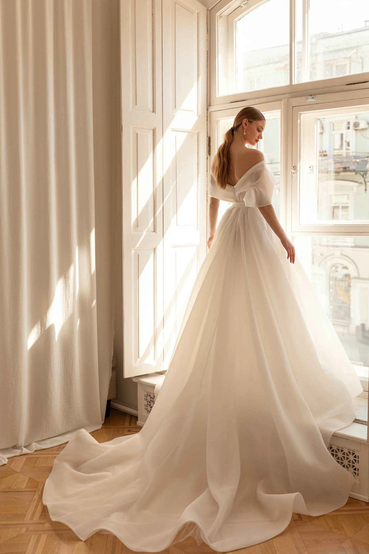 A woman wearing a voluminous off-the-shoulder wedding gown stands by a sunlit window, with the long train pooling on a wood floor in a bright interior.
