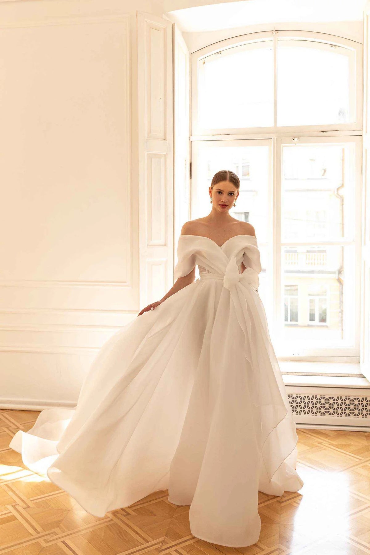 A woman wearing a voluminous off-the-shoulder white wedding gown stands in a bright room with large windows and wood floors, holding out the skirt to show its full shape.